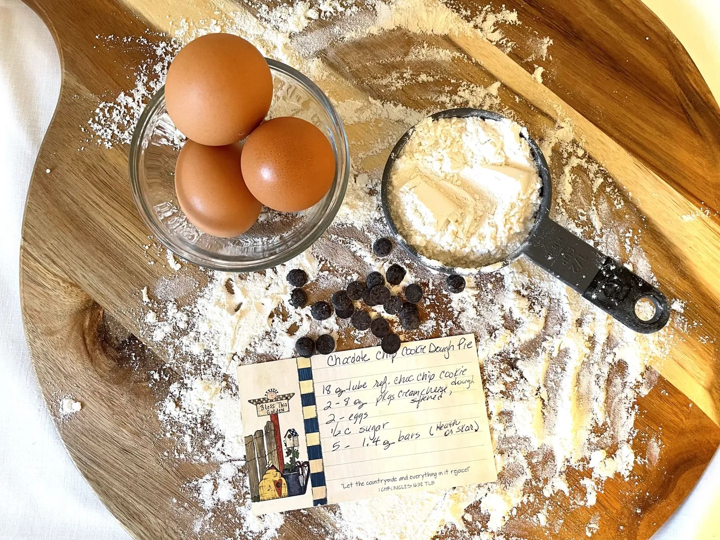 Three brown eggs in a glass bowl, a measuring cup of flour and a handwritten recipe card on a floured wooden board.