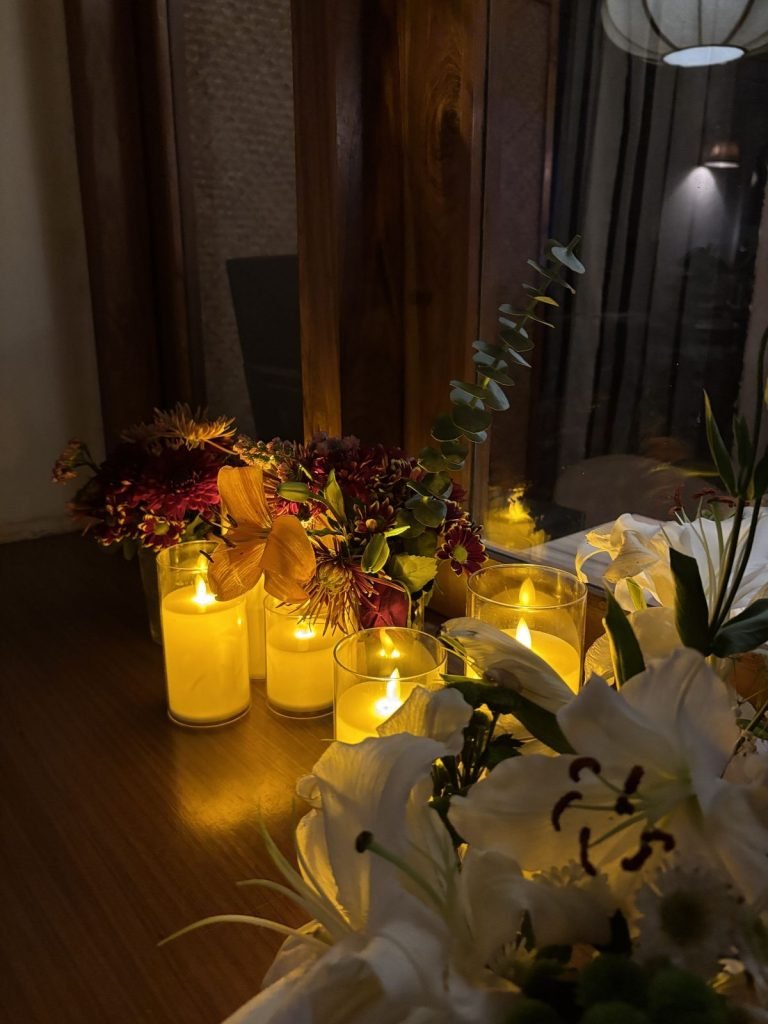 Lit candles and a vase of lilies and autumn flowers on a table beside a window in the evening.