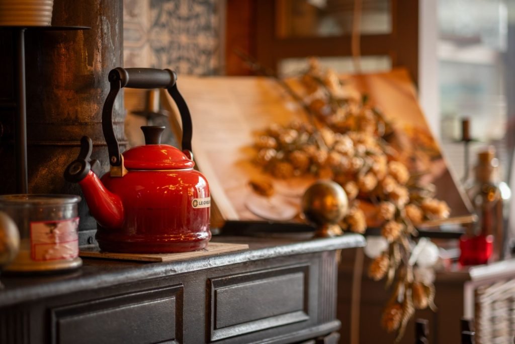 A red enamel kettle on a dark wooden cupboard in a kitchen, with dried autumn decorations behind it.