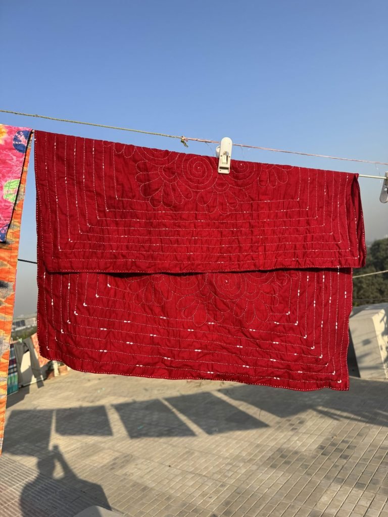 A deep red hand-stitched quilt pinned to a clothesline on a sunny rooftop, its shadow falling on the tiles.