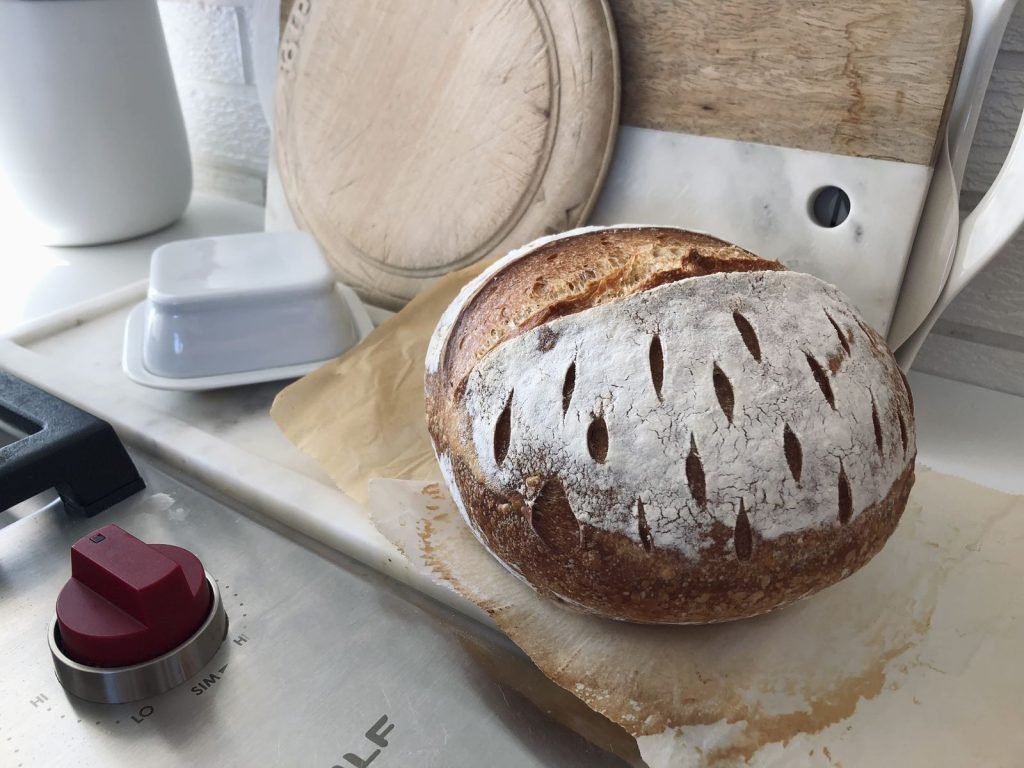 A scored round sourdough loaf cooling on a marble kitchen counter beside a bread board and a butter dish.