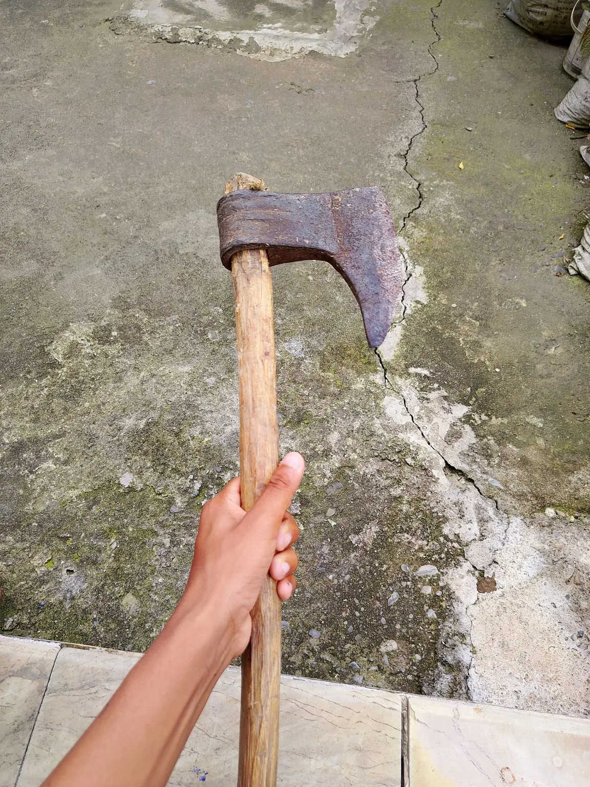 A hand holds an old axe with a rusty head and a wooden handle above a cracked, weathered concrete floor.
