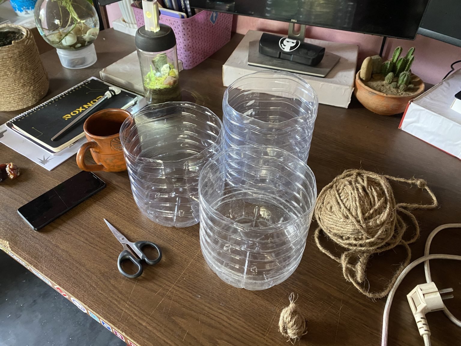 A wooden desk laid out with containers, twine, scissors, a phone, a mug, a notebook and small plants.
