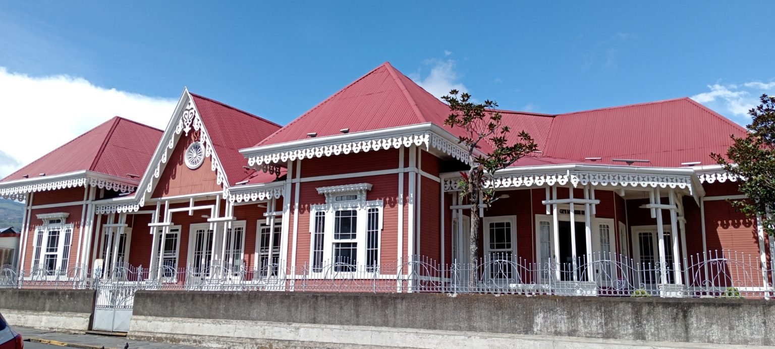 Close view of an ornate gable on a red Victorian-style building in Cartago, Costa Rica. White fretwork trim frames a round rosette window above double doors, with Libreria and Cafeteria signs marking a UNED university center.