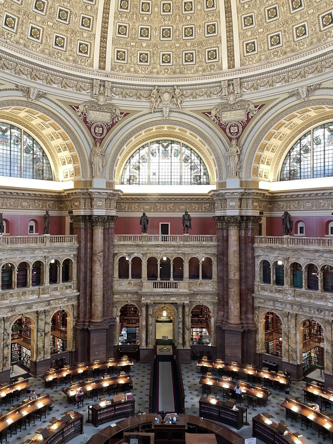 A domed library reading room with arched stained-glass windows, marble columns and rows of lit reading desks