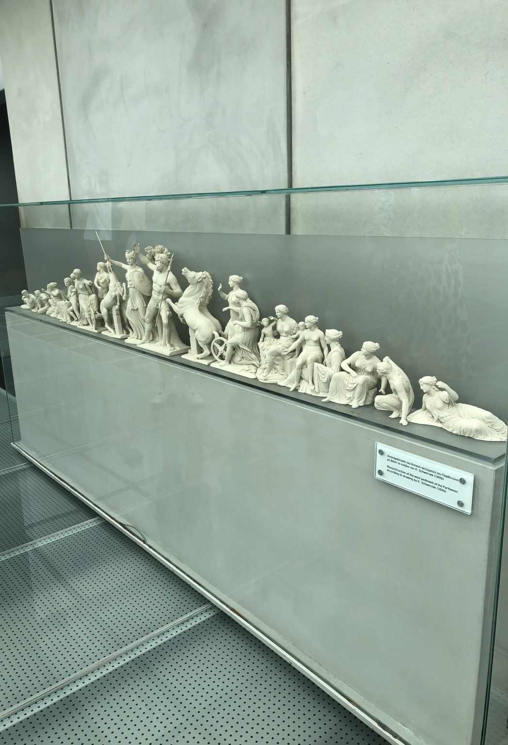 A long glass case holding a row of small white figurative sculptures, with a bilingual label on the plinth