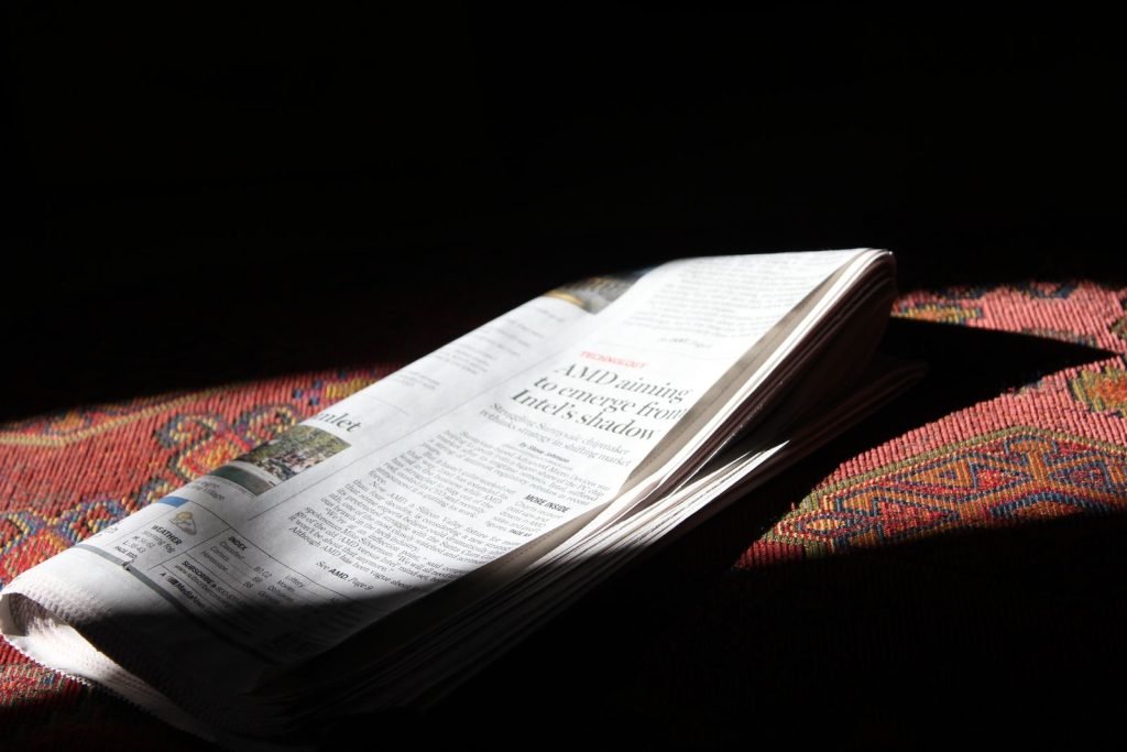 A folded newspaper lies in a strip of sunlight on a patterned red cloth.