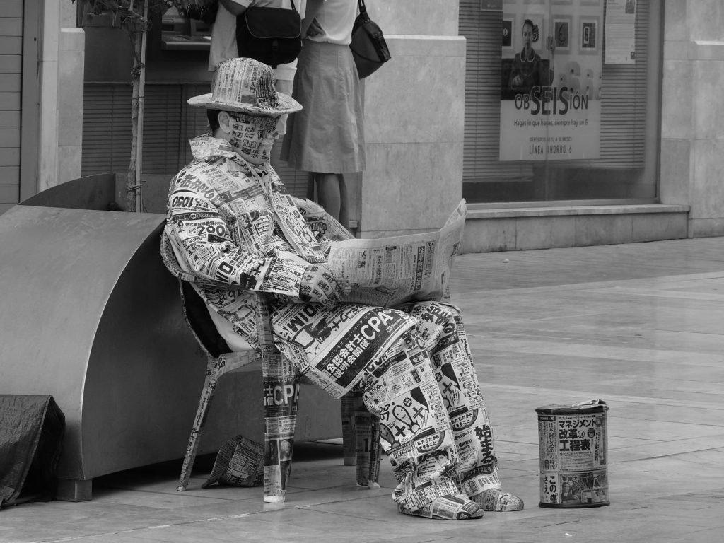 A street performer dressed entirely in newspaper sits on a chair reading a newspaper.