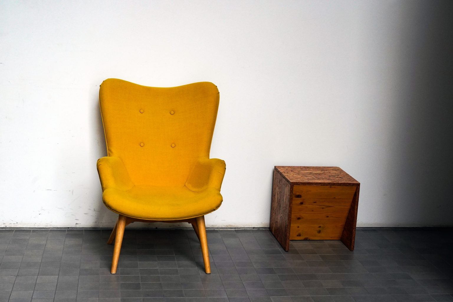 A mustard-yellow armchair and small wooden side table stand against a white wall.