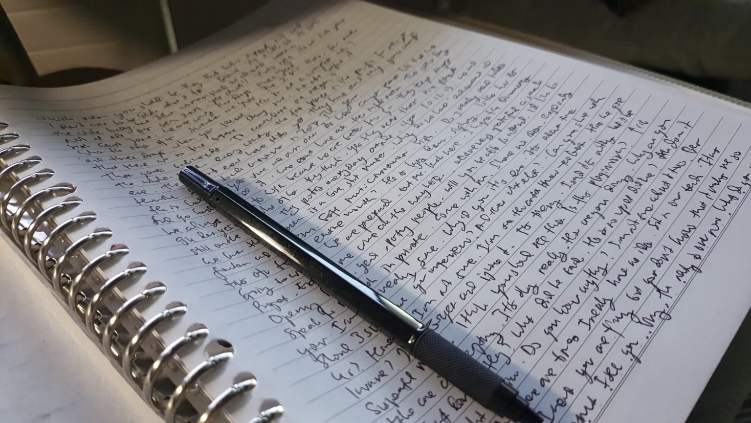 A black pen rests diagonally across a spiral notebook filled with handwriting.