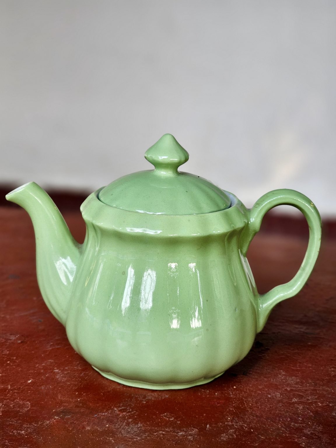 A pale green ceramic teapot stands on a red tabletop against a plain wall.