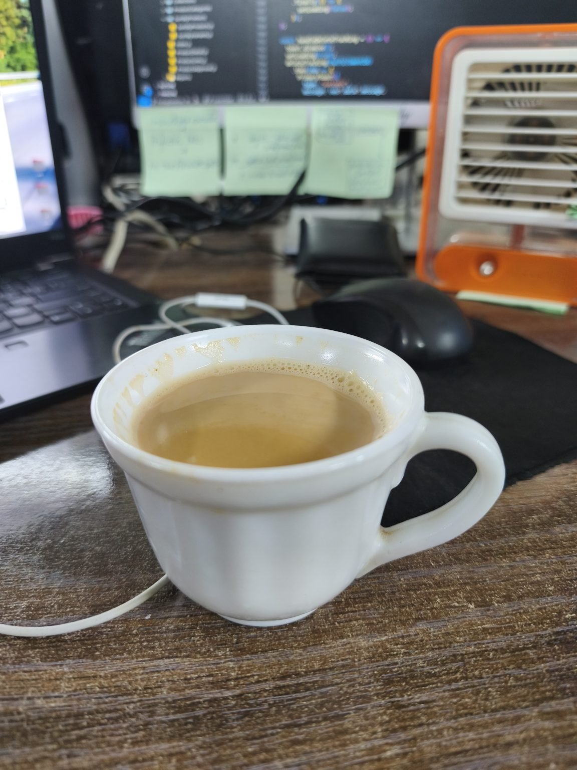 A white cup of tea sits on a wooden desk, surrounded by a laptop, sticky notes, and a small fan.