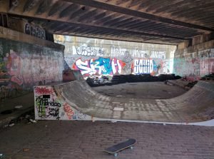 A skateboard rests in front of a worn ramp under a bridge covered in graffiti.