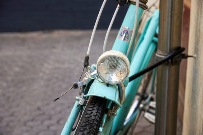 A weathered turquoise bicycle has a round headlamp above the front tire.
