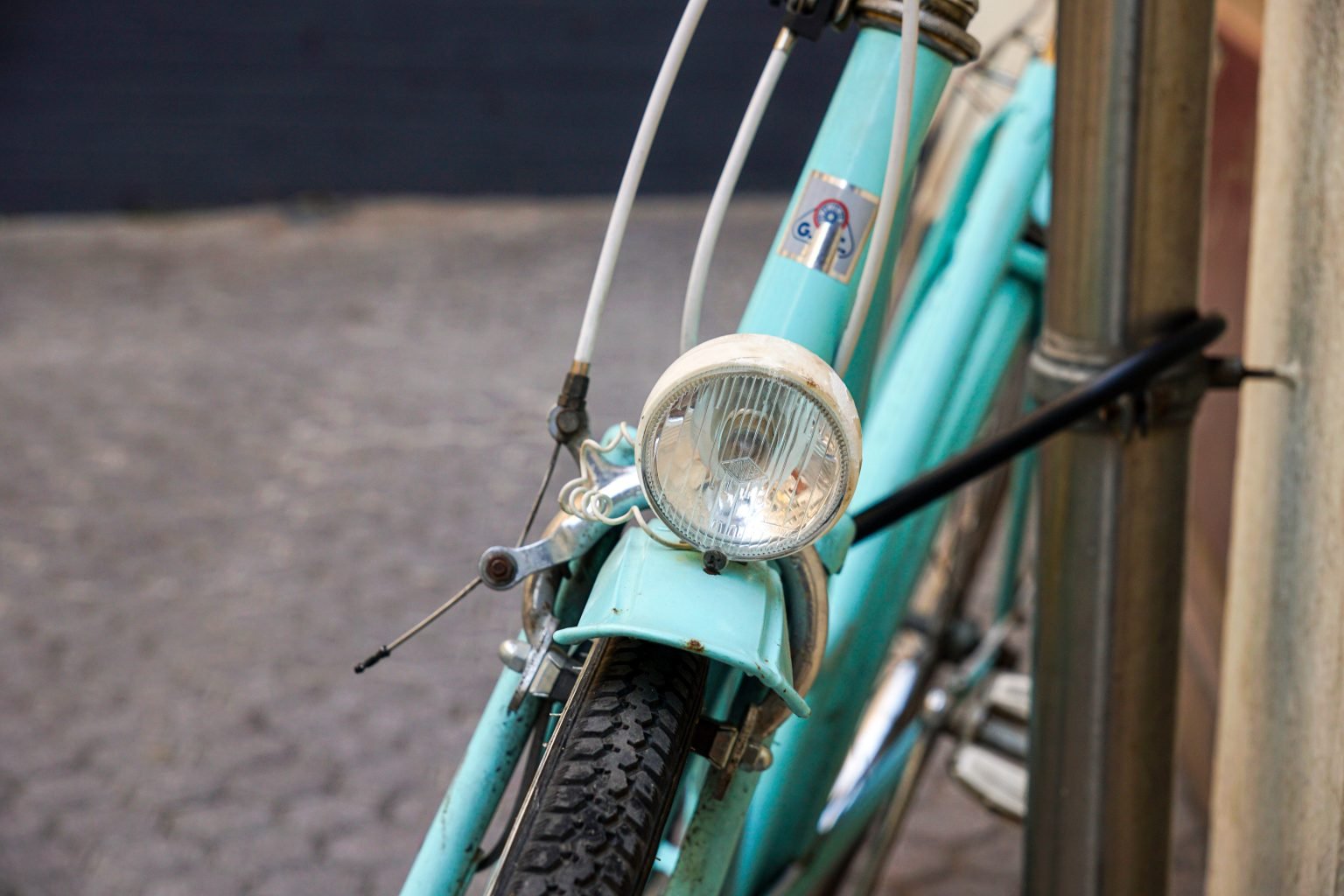 A weathered turquoise bicycle has a round headlamp above the front tire.
