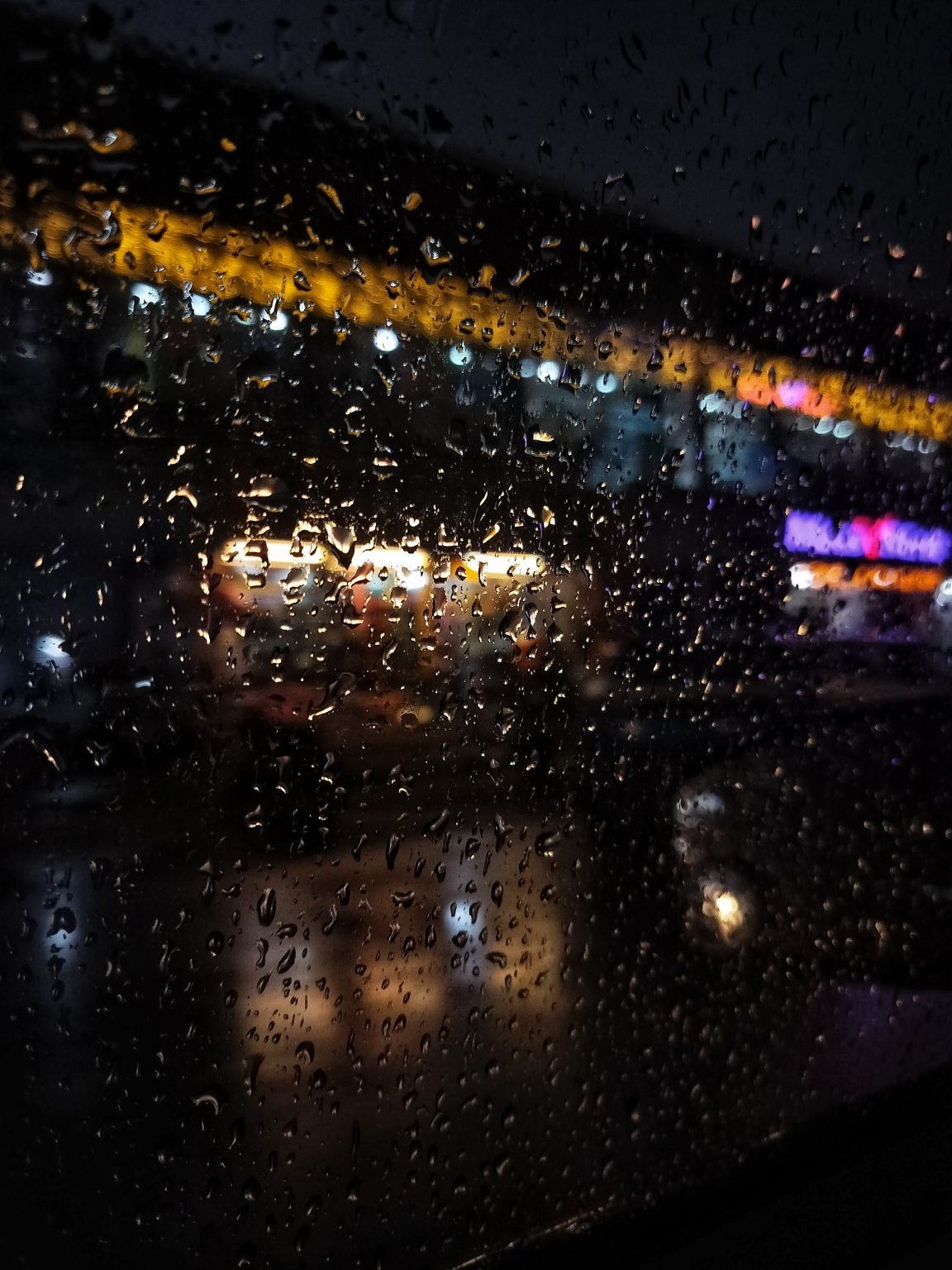 Rain droplets cover a window, with blurred street lights glowing beyond the glass.